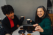 Positive ethnic mature woman having latte and taking self portrait with laughing African American girlfriend in outdoor coffee house