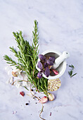 From above pestle and mortar with blossoming flowers near garlic cloves and fresh rosemary on marble table