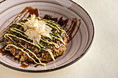 From above of tasty traditional Japanese okonomiyaki dish with sauce topped with bonito flakes served in ceramic bowl on light background