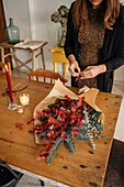 From above of crop unrecognizable female florist standing at wooden table with candles and arranging Christmas bouquet in room