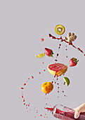 Bunch of various fresh fruits and ginger falling into glass bottle being hold by cropped anonymous person with healthy juice against gray background