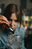 Soft focus of focused female barkeeper adding star anise into wineglass with alcoholic gin and tonic cocktail during work in bar