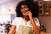 Glad young African American woman in apron and glasses leaning on counter and talking on cellphone while working in zero waste grocery store
