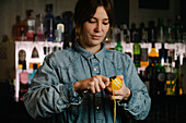 Focused female bartender taking off lemon zest while preparing for making cocktail in bar with various alcohol drinks during work