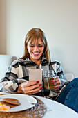 Smiling adult female in plaid shirt lounging on sofa at table with breakfast meal and having drink while browsing social media on phone