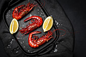 Delicious cooked red prawns on tray with coarse salt and juicy lemon pieces on dark background