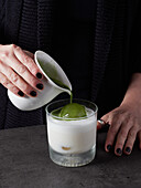 Anonymous female pouring aromatic matcha beverage into glass cup with milk and ice cube while preparing healthy drink