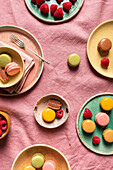 Ansicht von oben auf verschiedene runde Teller mit verschiedenen süßen, bunten französischen Makronen und Beeren auf rosa Tischtuch