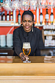 Cheerful black female barkeeper serving glass of refreshing beer in bar looking at camera