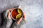 Kid hands in mittens hold cup of tea with lemon on grey concrete background