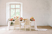 Stühle auf einem Tisch mit weißer Tischdecke und Blumen vor einem Fenster in einem sonnenbeschienenen Raum mit schäbigen Wänden während einer Hochzeitsfeier