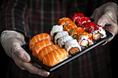 Crop unrecognizable chef in gloves showing platter with set of palatable assorted sushi in dark room