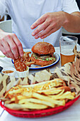 Crop anonymous man in white t shirt sitting at table with appetizing burger hot dog French fries and beer while dipping crouton in sauce