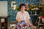 Happy female florist creating wreath from fresh colorful flowers while working at table in shop