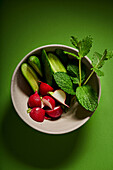 From above of cut fresh cucumbers with radish and leaves of mint placed in bowl served on green table