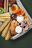 From above brunch box with assorted sliced meats various types of cheese and crispbreads arranged near ripe cup kiwi sweet strawberries and peeled mandarin near jam in glass jar on colorful green background