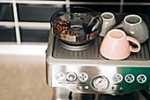 Blick von oben auf geröstete Kaffeebohnen in einer elektrischen Kaffeemühle und Keramiktassen auf einem Gestell einer modernen Espressomaschine zu Hause