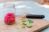 Green herbs and lime pieces placed near vegetable peeler and glass jar with pomegranate seeds on wooden chopping board