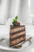 Piece of yummy sweet chocolate cake decorated with cream served on ceramic plate with golden spoon on white tablecloth