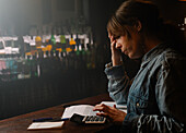 Side view of focused female worker using calculator to count income while sitting at counter with documents during work in cafe