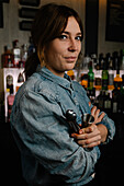 Attractive female bartender with jigger and tong in hands looking at camera while standing in bar with various alcohol drinks