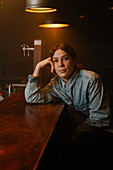 Calm female in denim shirt leaning on hand and looking at camera while sitting at wooden counter during work in bar