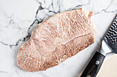 Top view of traditional Japanese uncooked kobe prefecture wagyu beef placed on marble table near knife in light room during cooking preparation