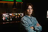 Attractive waitress in apron looking at camera with crossed arms while standing near bar counter with bottles of alcohol drinks
