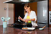Cheerful adult woman pouring batter on hot electric griddle while cooking pancakes at counter in modern kitchen