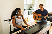 Ruhige hispanische Frau mit Kopfhörern, die E-Piano spielt und in ein Mikrofon singt, während sie mit einem Freund, der Akustikgitarre spielt, tagsüber zu Hause Musik macht