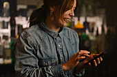 Positive female barkeeper surfing internet on cellphone while standing in bar with various alcoholic drinks during work in modern cafe