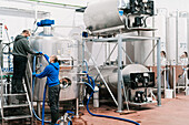 Side view of male coworkers in face masks connecting hose to metal reservoir in beer factory during coronavirus pandemic