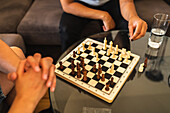 From above of crop unrecognizable friends in casual clothes sitting at glass table and playing chess in room in daytime