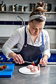 Woman in apron using tweezers to put slice of fresh strawberry on top of whipped cream while cooking dessert in kitchen of contemporary restaurant