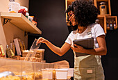 Low angle of young black female vendor in uniform and glasses with tablet smiling and inspecting pasta in glass container during work in zero waste grocery store