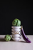 Ripe green artichokes and eggplant with precious accessories hanging on stand placed on table against black background