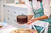 Crop unrecognizable female in apron standing near table while demonstrating tasty cake with chocolate cream