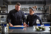 Plump man showing plate with pork slices to female colleague in black uniform during work in kitchen of modern restaurant