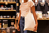 Cropped unrecognizable young African American woman in casual clothes with curly hair smiling and using tote bag while visiting eco friendly grocery shop