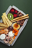 From above brunch box with assorted sliced meats various types of cheese and crispbreads arranged near ripe cup kiwi sweet strawberries and peeled mandarin near jam in glass jar on colorful green background