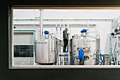 Side view of male coworkers in face masks connecting hose to metal reservoir in beer factory during coronavirus pandemic