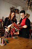Fröhliche Freundinnen stehen am Tisch mit Kerzen und basteln kreative Weihnachtssträuße für die Weihnachtsfeier