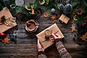 Top view of an anonymous woman preparing a Christmas present next to a bowl of chocolate and Christmas decorations on a wooden table