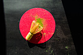 Ansicht von oben auf einen schwarzen Tisch im Sonnenlicht mit Bambus und getrocknetem Matcha-Pulver auf einem rosa Kreis