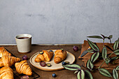 Tasty fresh baked croissants served on plate with fruits placed near cup of tea on wooden table in morning time in light room