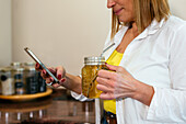 Cropped unrecognizable smiling adult female in casual outfit leaning on counter in kitchen browsing smartphone and holding glass mug of refreshing beverage