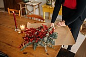 From above of crop unrecognizable female florist standing at wooden table with candles and arranging Christmas bouquet in room