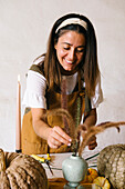 Positive adult Hispanic female designer with long dark hair in apron smiling while decorating table with reed plants pumpkins and candles for Halloween celebration