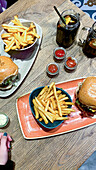 High angle of burgers with French fries placed on plates near ketchup jars and cold tea in glass cups with lemon and straw in cafe