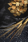 Pieces of crispy bread in bowl near wheat spikes on black textile in room